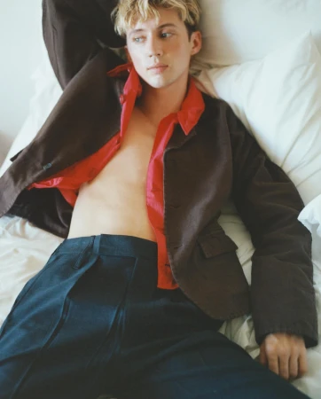 Young adult with blond hair poses in bed, wearing a brown jacket and blue pants, showcasing fashion and lifestyle elements.