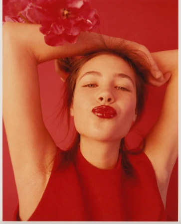 Red toned portrait of a young adult woman with a flower, hands behind her head and lips puckered against a solid red backdrop.
