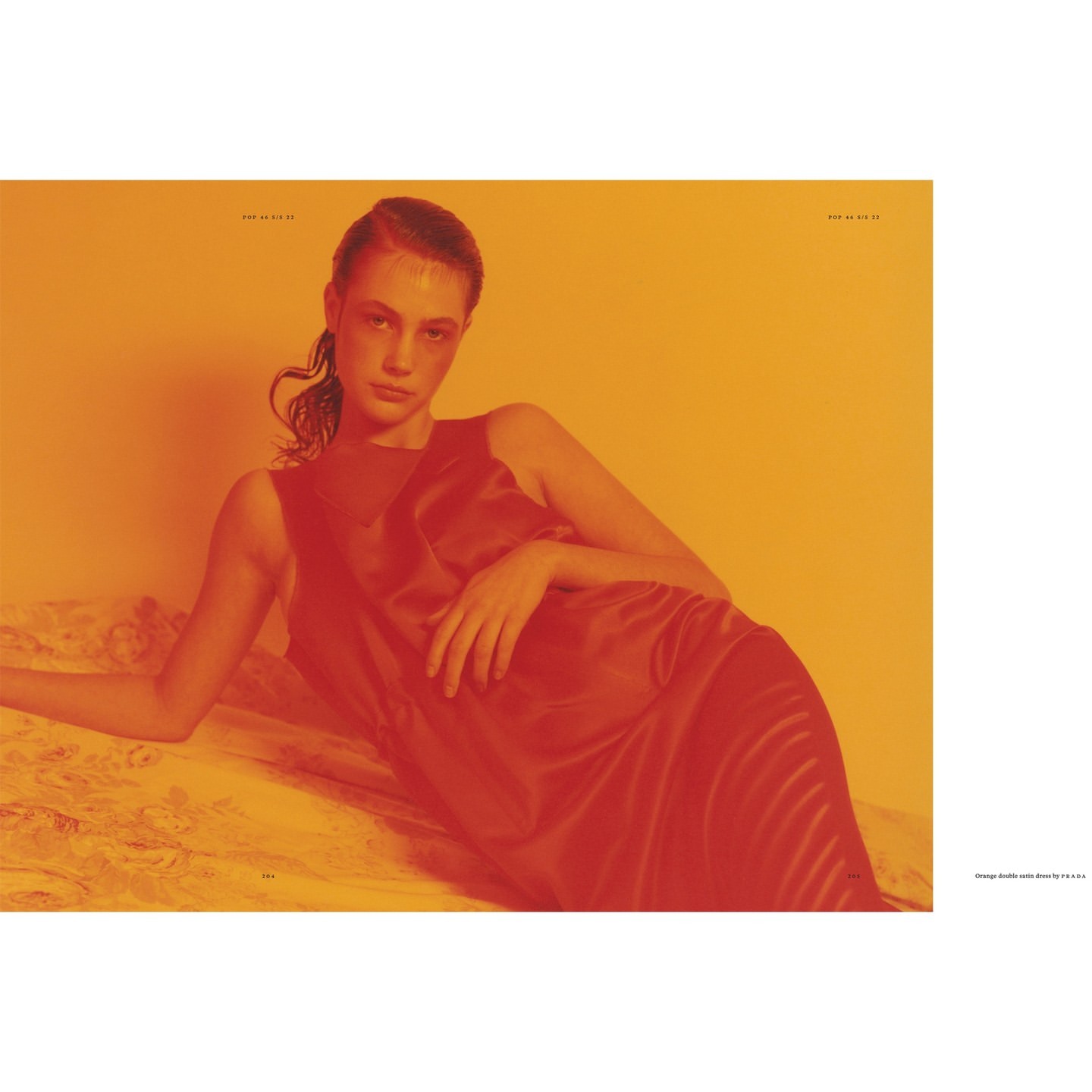 Woman reclines in a red sleeveless dress on a textured surface, set against an orange background in a fashion or beauty editorial.