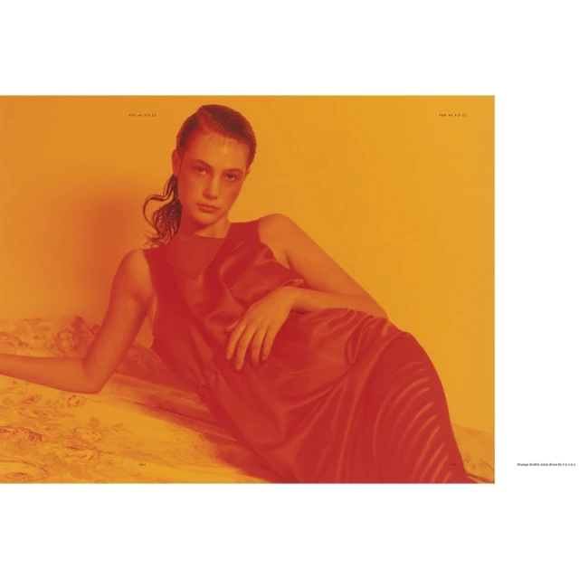 Woman reclines in a red sleeveless dress on a textured surface, set against an orange background in a fashion or beauty editorial.