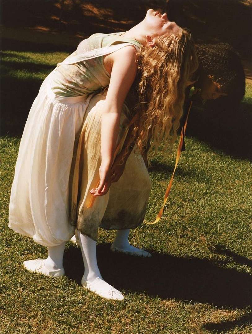 Woman with long blonde hair and light skin wears a white dress and white tights, bending backward in a green grassy outdoor setting.