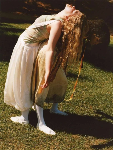 Woman with long blonde hair and light skin wears a white dress and white tights, bending backward in a green grassy outdoor setting.