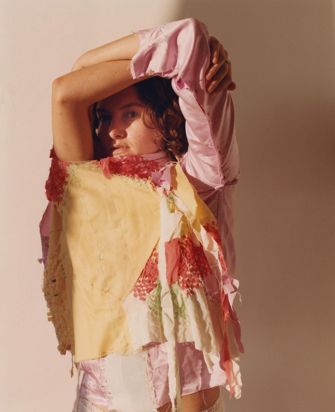 Portrait of a female with arms raised behind her head, wearing a patchwork top in a photo studio with neutral lighting and a white backdrop.
