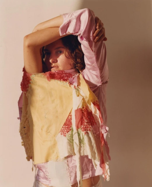 Portrait of a female with arms raised behind her head, wearing a patchwork top in a photo studio with neutral lighting and a white backdrop.