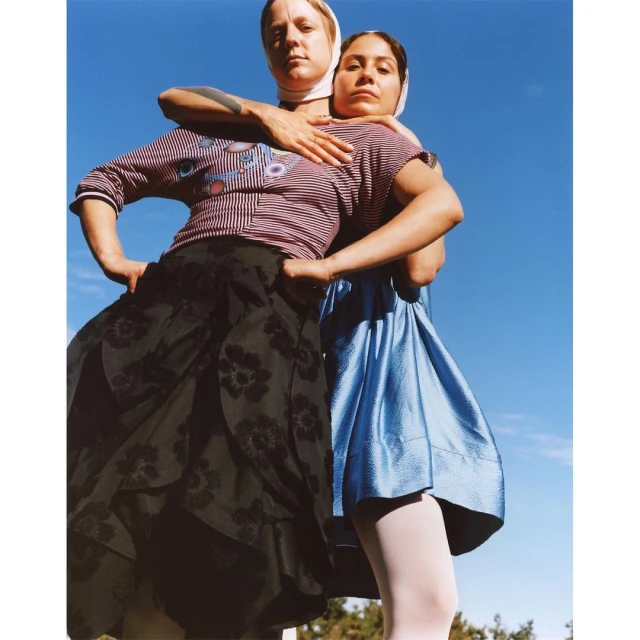 Two women in skirts pose outdoors against a blue sky backdrop. One woman has her arms around the other's shoulders in a friendly way.