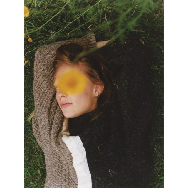 Woman in a dark sweater and a light-brown sweater, lying in the grass with yellow flowers in the background, outdoor portrait.