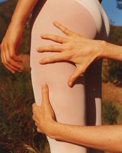 Close-up of a person's thigh and hip in white tights, with two hands touching the body, set against an outdoor, leafy background.