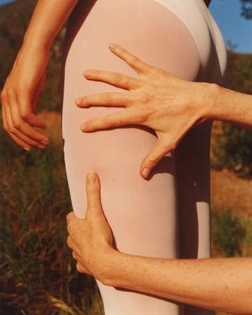 Close-up of a person's thigh and hip in white tights, with two hands touching the body, set against an outdoor, leafy background.