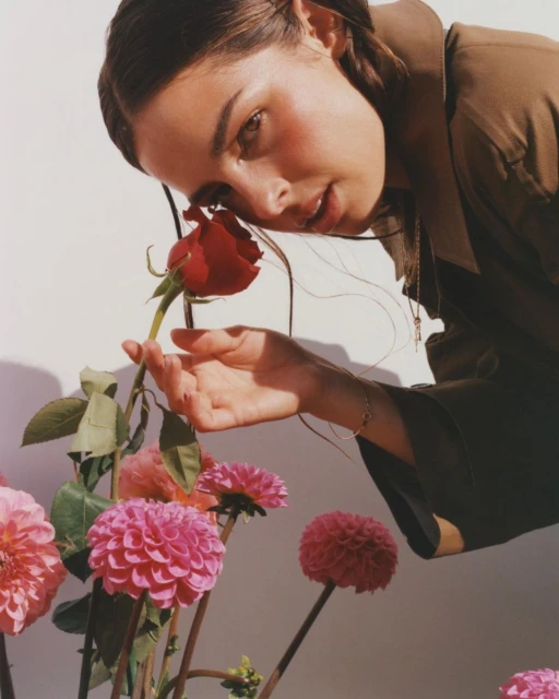 Close-up of a brunette woman in a brown jacket, holding a red rose, surrounded by pink dahlia flowers against a neutral background.
