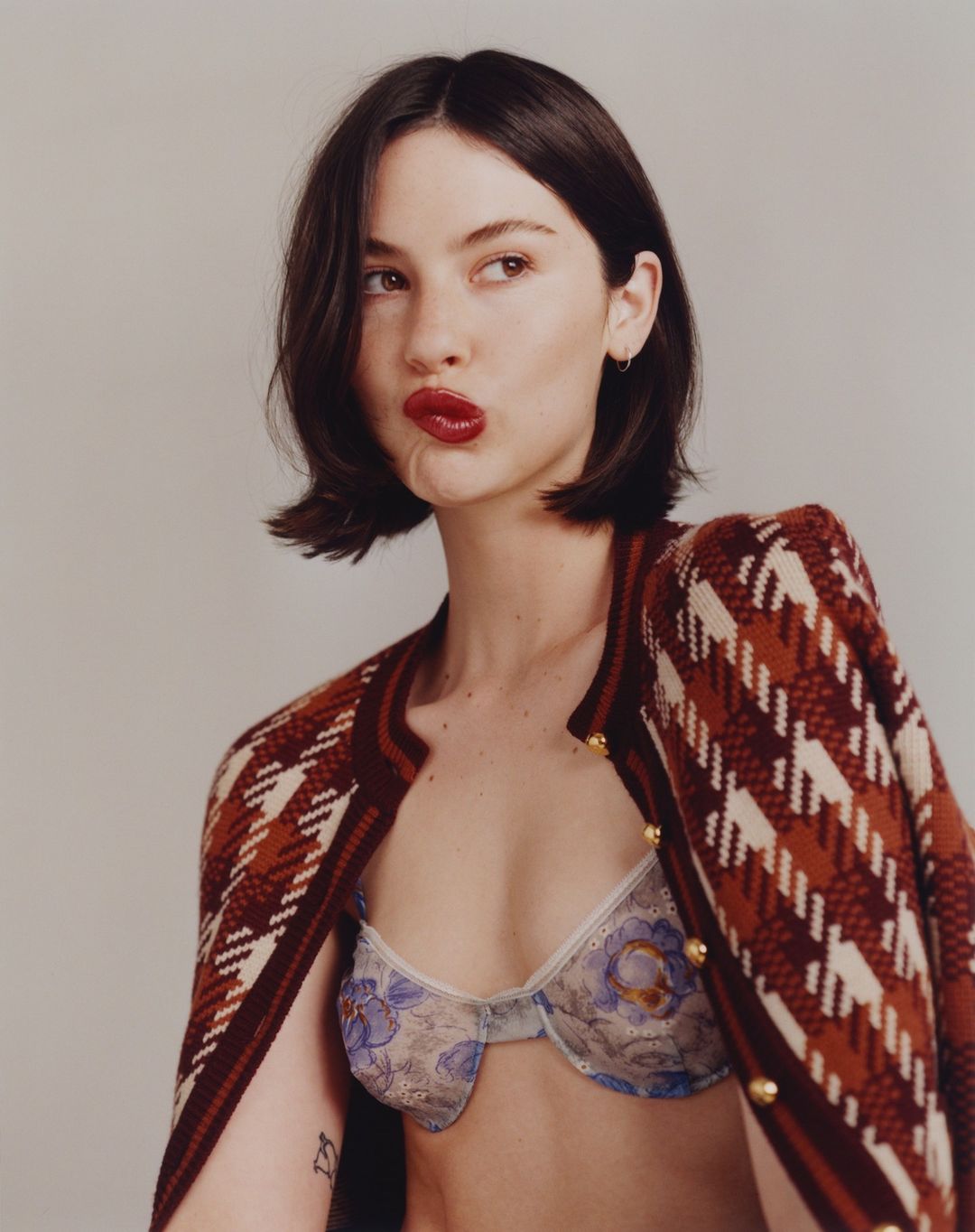 Portrait of a woman with dark hair wearing an open cardigan and bra, pouting her lips, shot against a neutral background.