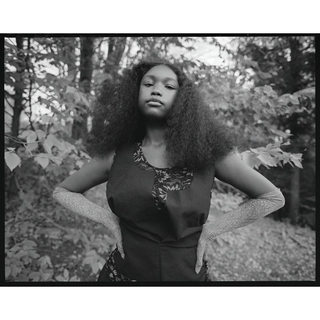 Monochromatic portrait of a young Black woman with curly hair, wearing a sleeveless dress and textured arm coverings, posing outdoors.