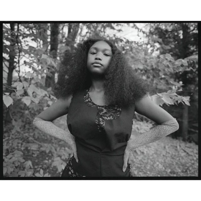 Monochromatic portrait of a young Black woman with curly hair, wearing a sleeveless dress and textured arm coverings, posing outdoors.