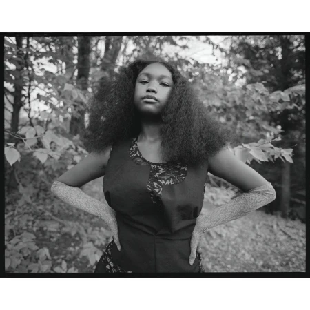 Monochromatic portrait of a young Black woman with curly hair, wearing a sleeveless dress and textured arm coverings, posing outdoors.