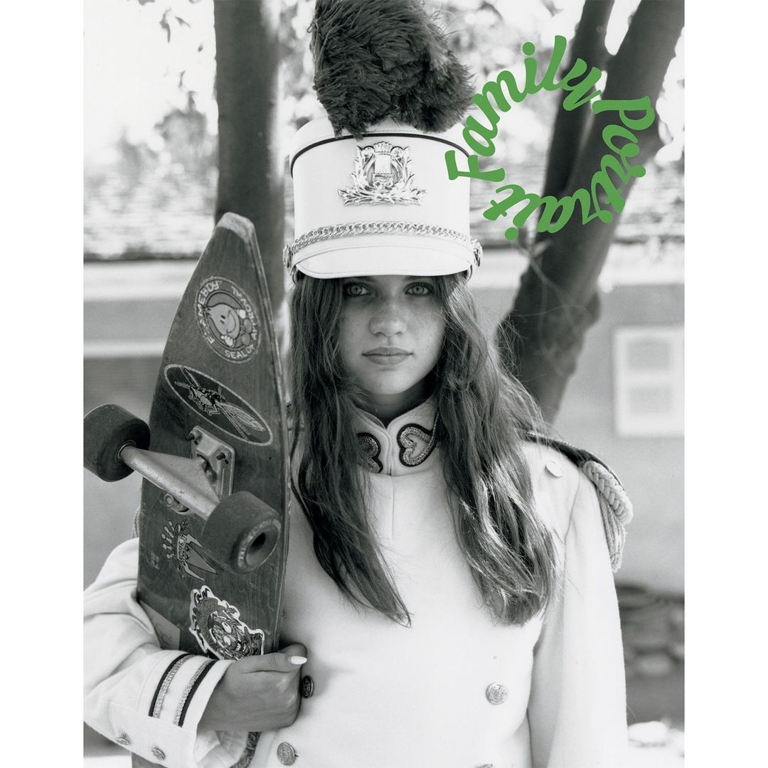 A young woman in a marching band uniform holds a skateboard in a monochromatic portrait with soft lighting and a blurred background.