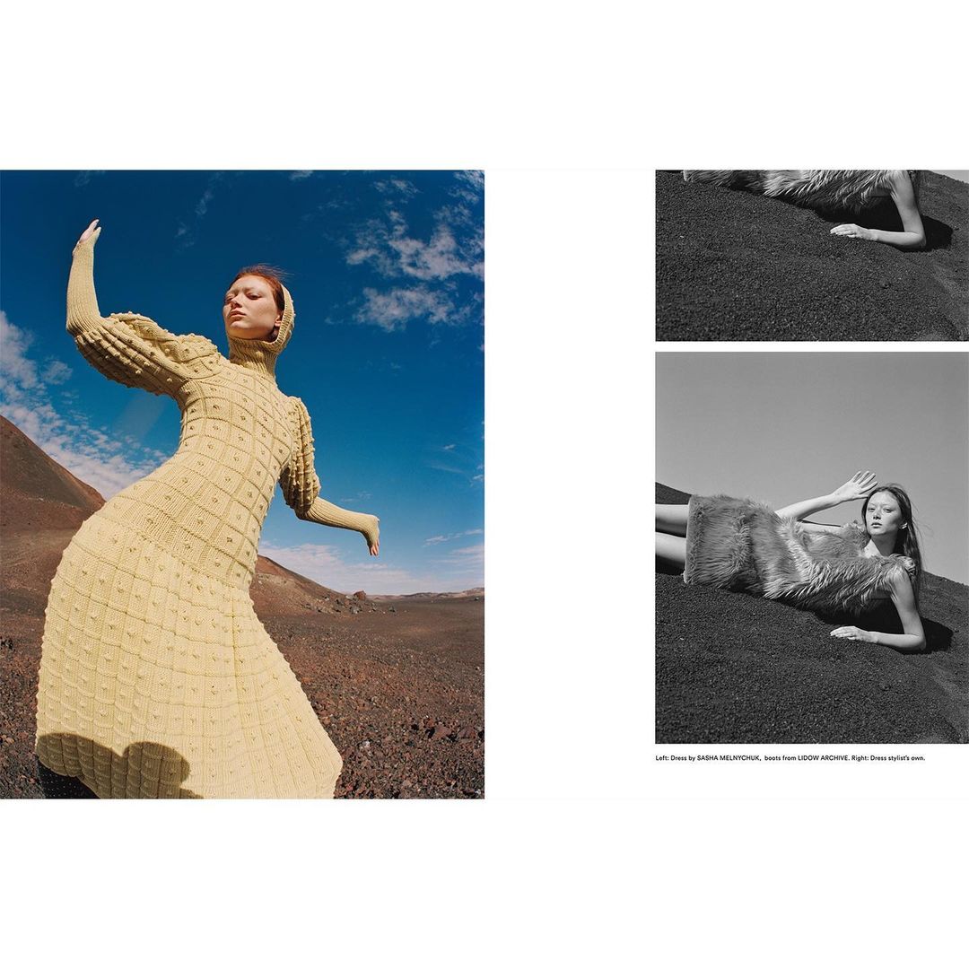 A woman in a light yellow dress with long sleeves and a head covering poses in an open desert landscape under a blue sky.