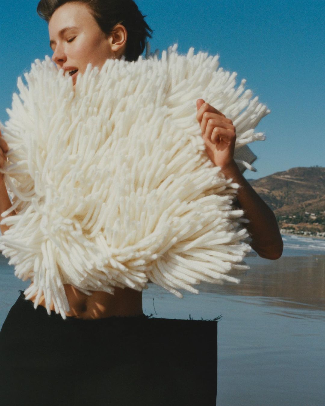 Woman in a textured white fringe garment standing on a sandy beach against an ocean backdrop