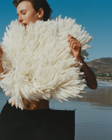 Woman in a textured white fringe garment standing on a sandy beach against an ocean backdrop