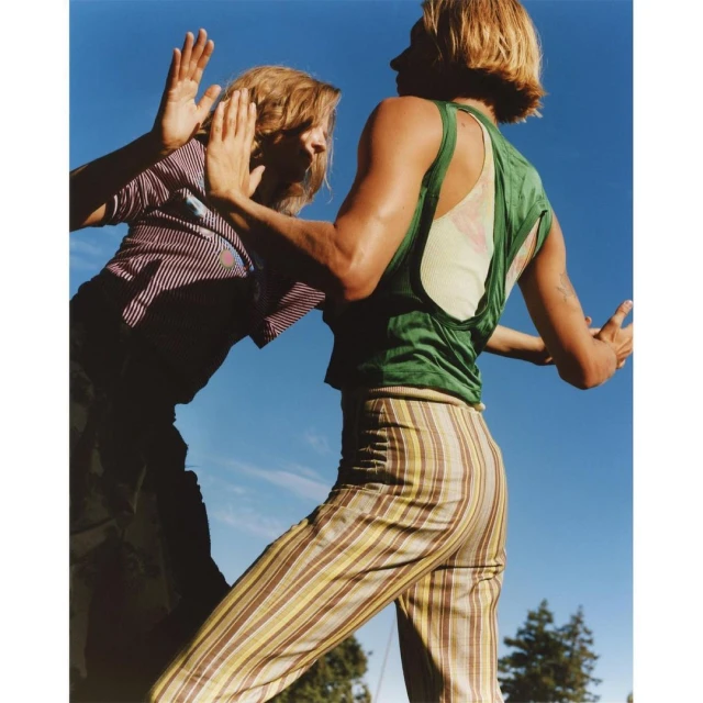 Two people face each other outdoors under a bright blue sky. One person raises their hands towards the other, who wears a green top.