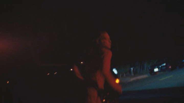 Woman with blonde hair walking at night on a road near parked cars, illuminated by warm-toned street lights and car headlights.
