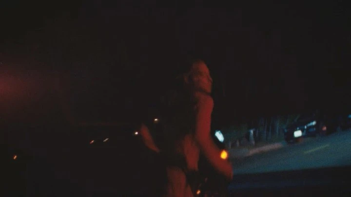Woman with blonde hair walking at night on a road near parked cars, illuminated by warm-toned street lights and car headlights.