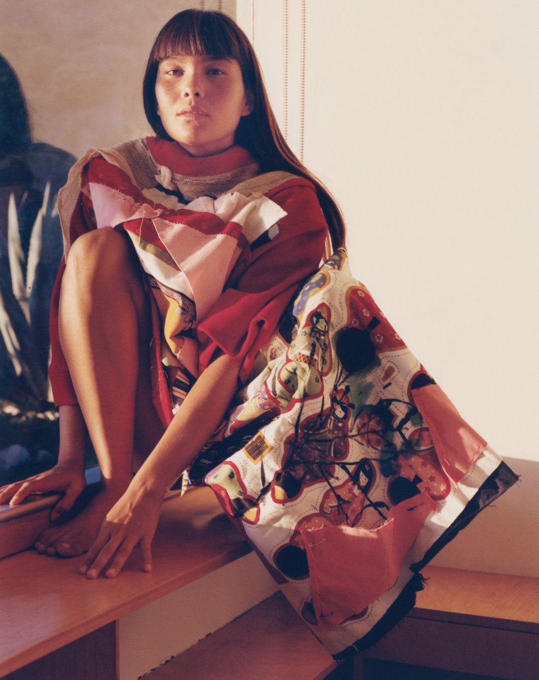Woman in a colorful patchwork-style garment sits on a ledge in a brightly lit room, featuring a blurred reflection in the background.