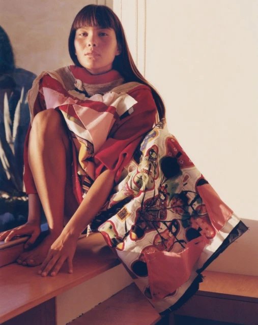Woman in a colorful patchwork-style garment sits on a ledge in a brightly lit room, featuring a blurred reflection in the background.