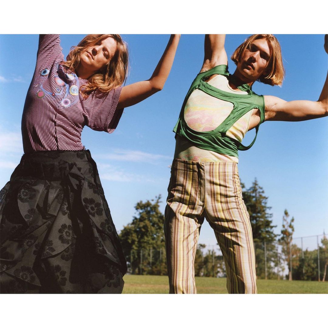 Two people pose outdoors in a field, dressed in casual, patterned clothing under a clear blue sky with scattered trees in background.