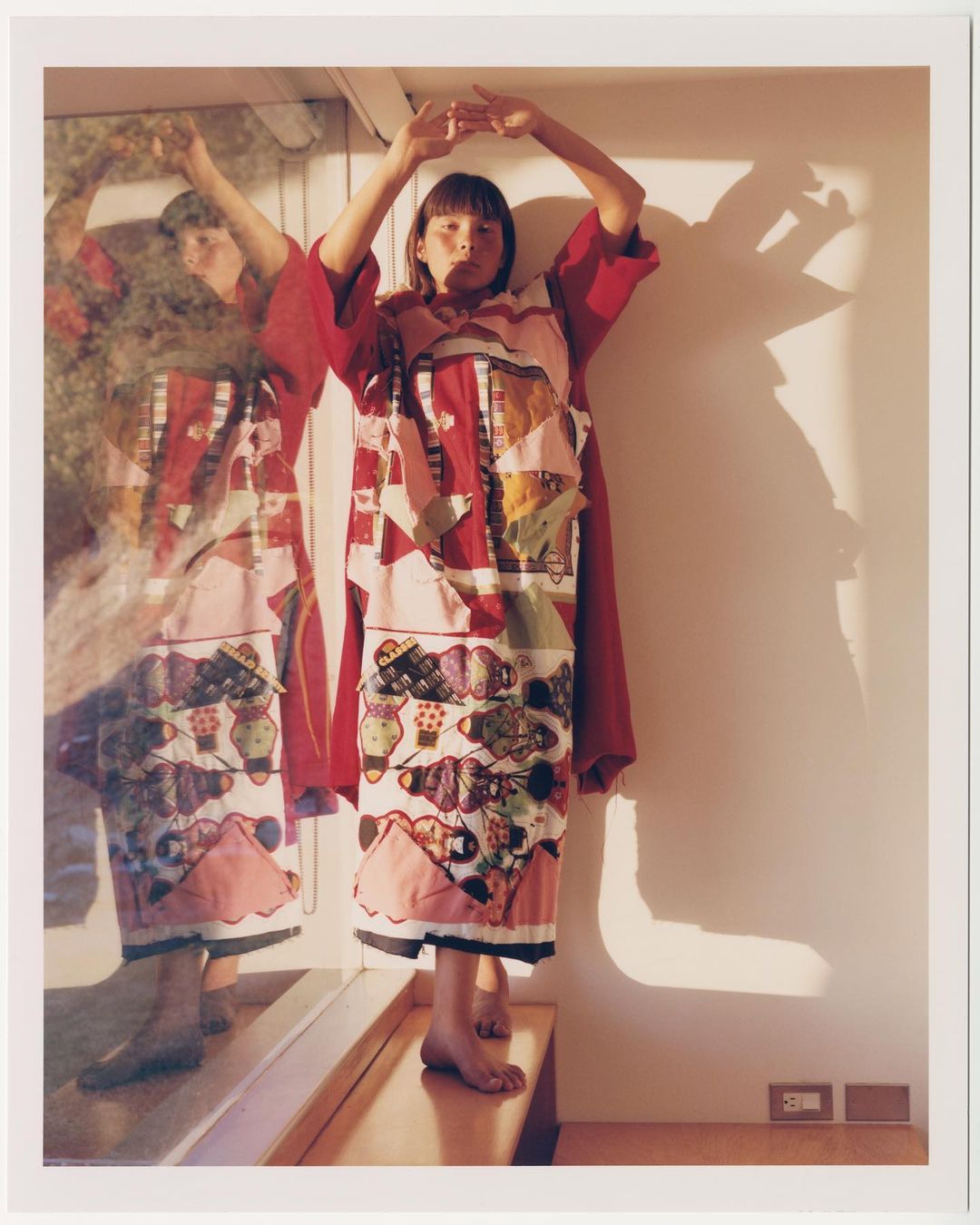 A young adult woman poses in a red and white patchwork dress in front of a mirror in an indoor setting, casting shadows on the wall.