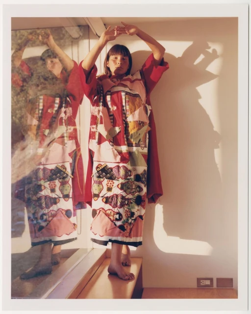 A young adult woman poses in a red and white patchwork dress in front of a mirror in an indoor setting, casting shadows on the wall.