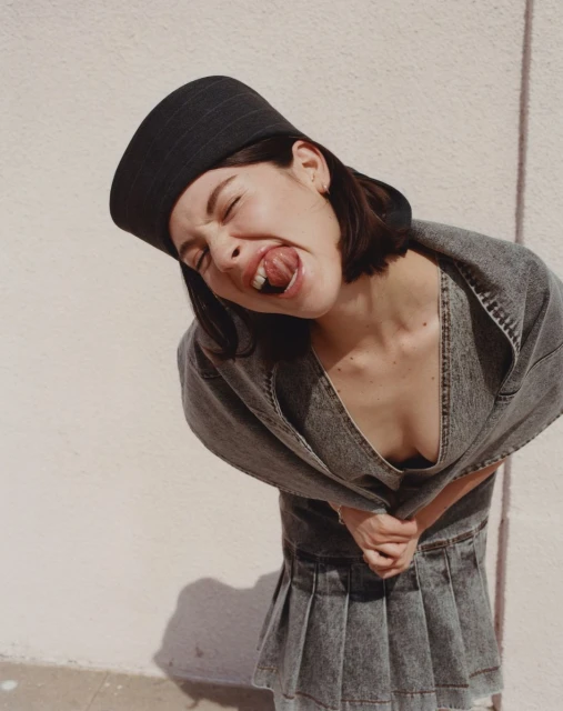 Woman in denim outfit with tongue out and eyes closed, wearing black headwear, poses outdoors against a light-colored wall in daylight.