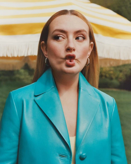 Headshot of a young adult female with auburn hair, wearing a turquoise blazer, pursing her lips, outdoors with greenery backdrop.