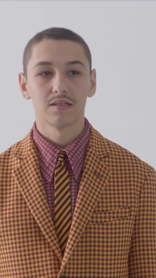 Young adult male in a yellow and brown checked suit with a striped tie stands indoors against a white background, portrait orientation.