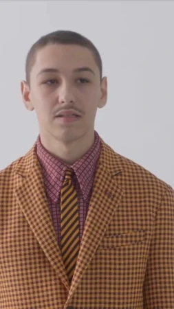Young adult male in a yellow and brown checked suit with a striped tie stands indoors against a white background, portrait orientation.