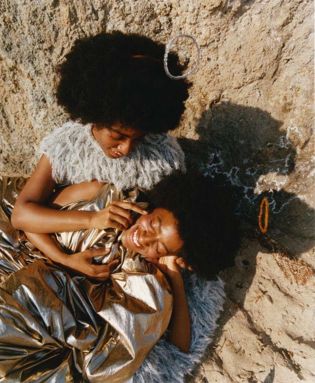 Two smiling Black women in a desert setting; one in gold fabric, the other in a fringed top, rocks, and strong light create a visually striking scene.