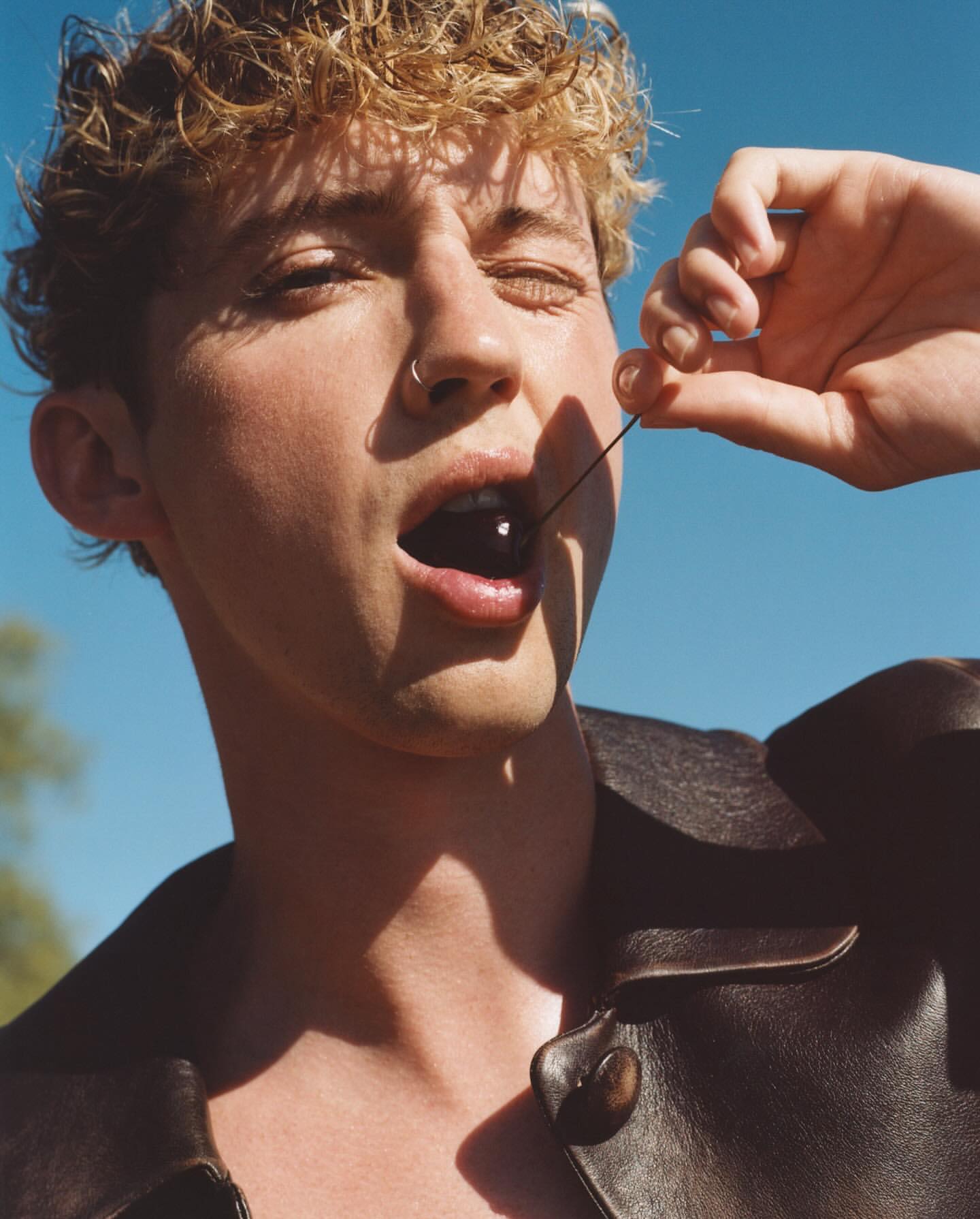 Close-up portrait of a young adult man eating a cherry on a blue sky backdrop. He is wearing a dark leather jacket.