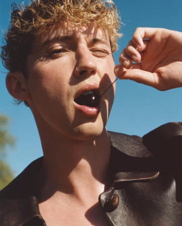 Close-up portrait of a young adult man eating a cherry on a blue sky backdrop. He is wearing a dark leather jacket.