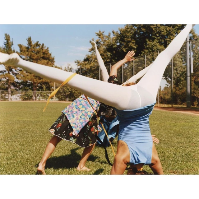 Two women in white tights and colorful tops doing handstands on a grass field, set against a backdrop of green trees and a clear blue sky.