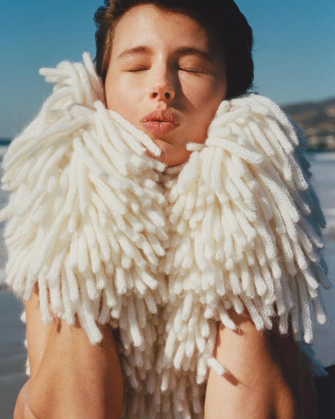 Portrait of a woman wearing a textured white fringe sweater with her eyes closed against an outdoor coastal background