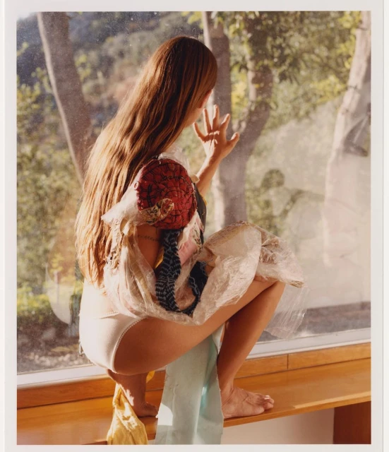 A woman with long brown hair sits crouched on a ledge by a window, partially dressed with trees visible outside.