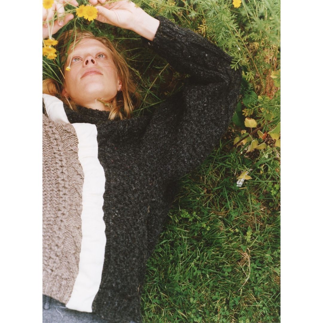 Fair-skinned adult woman lying in green grass with yellow flowers wearing a grey sweater. Relaxed, elevated shot, eye-level.