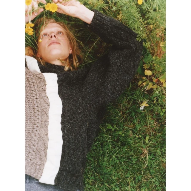 Fair-skinned adult woman lying in green grass with yellow flowers wearing a grey sweater. Relaxed, elevated shot, eye-level.