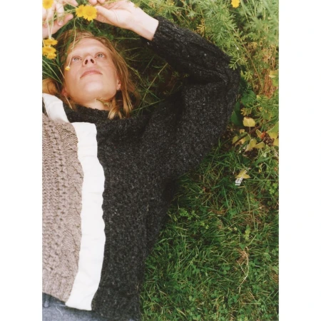 Fair-skinned adult woman lying in green grass with yellow flowers wearing a grey sweater. Relaxed, elevated shot, eye-level.