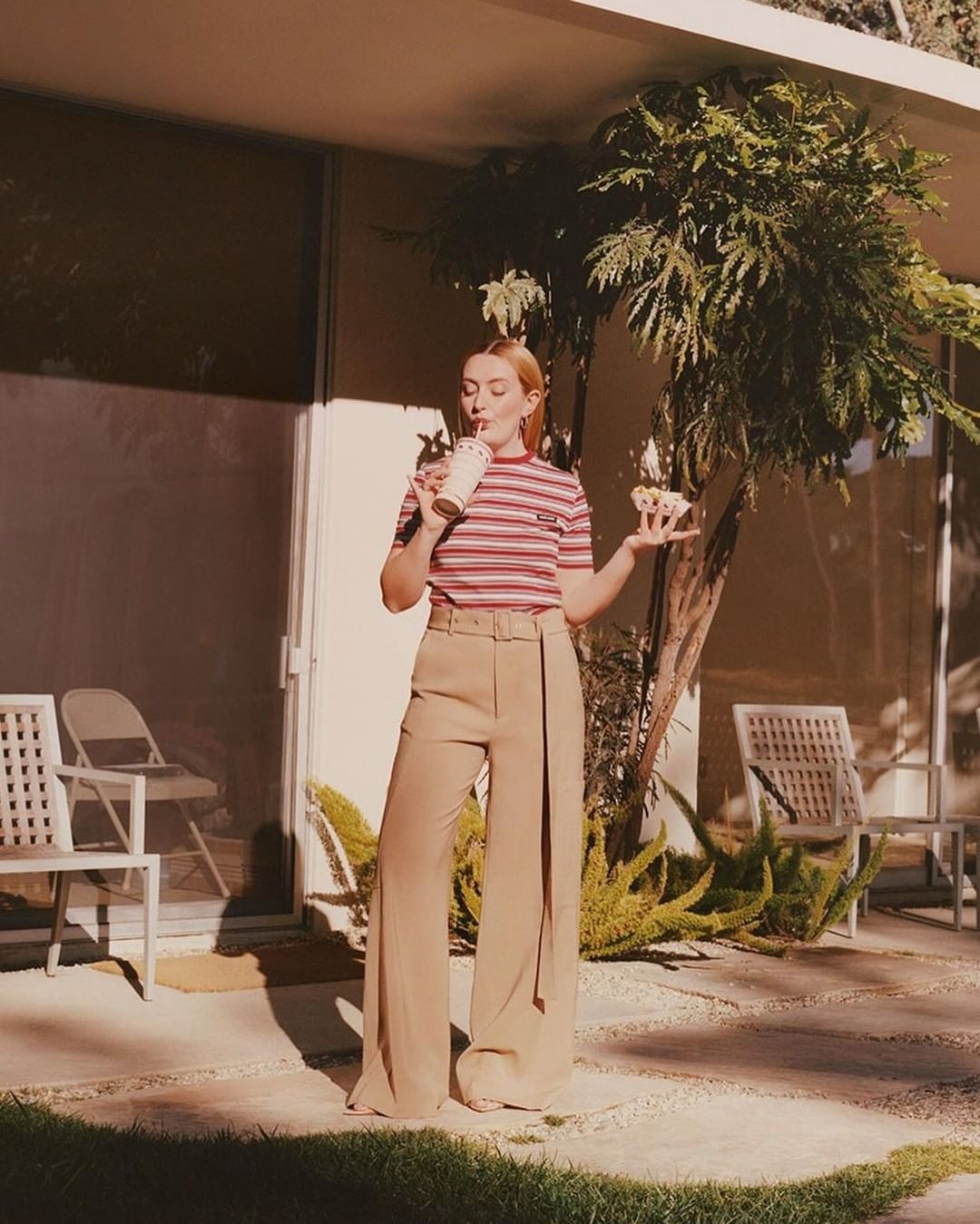 Woman in striped top and beige pants drinking from a cup and holding pastry outdoors in a sunlit backyard with white chairs and greenery.