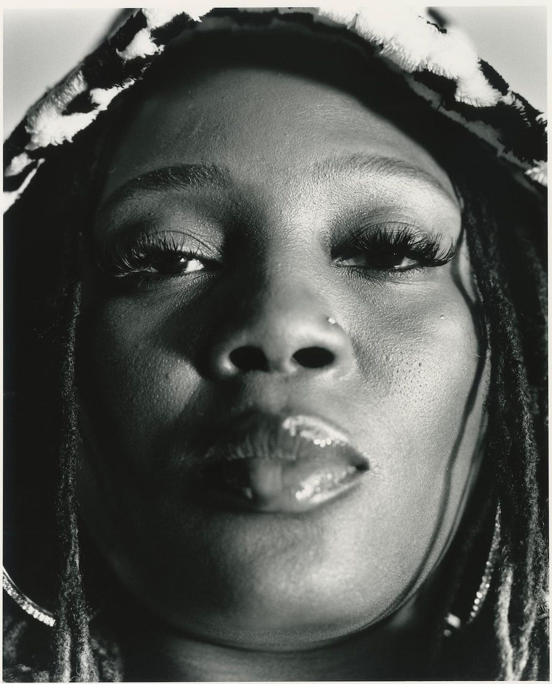 Close-up black-and-white portrait of an adult woman with dreadlocks wearing a patterned hood, focusing on her face, nose ring, and glossy lips.