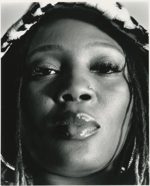 Close-up black-and-white portrait of an adult woman with dreadlocks wearing a patterned hood, focusing on her face, nose ring, and glossy lips.