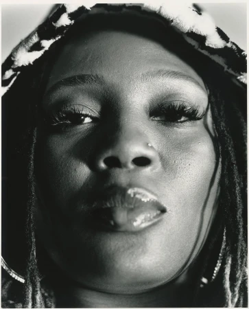 Close-up black-and-white portrait of an adult woman with dreadlocks wearing a patterned hood, focusing on her face, nose ring, and glossy lips.