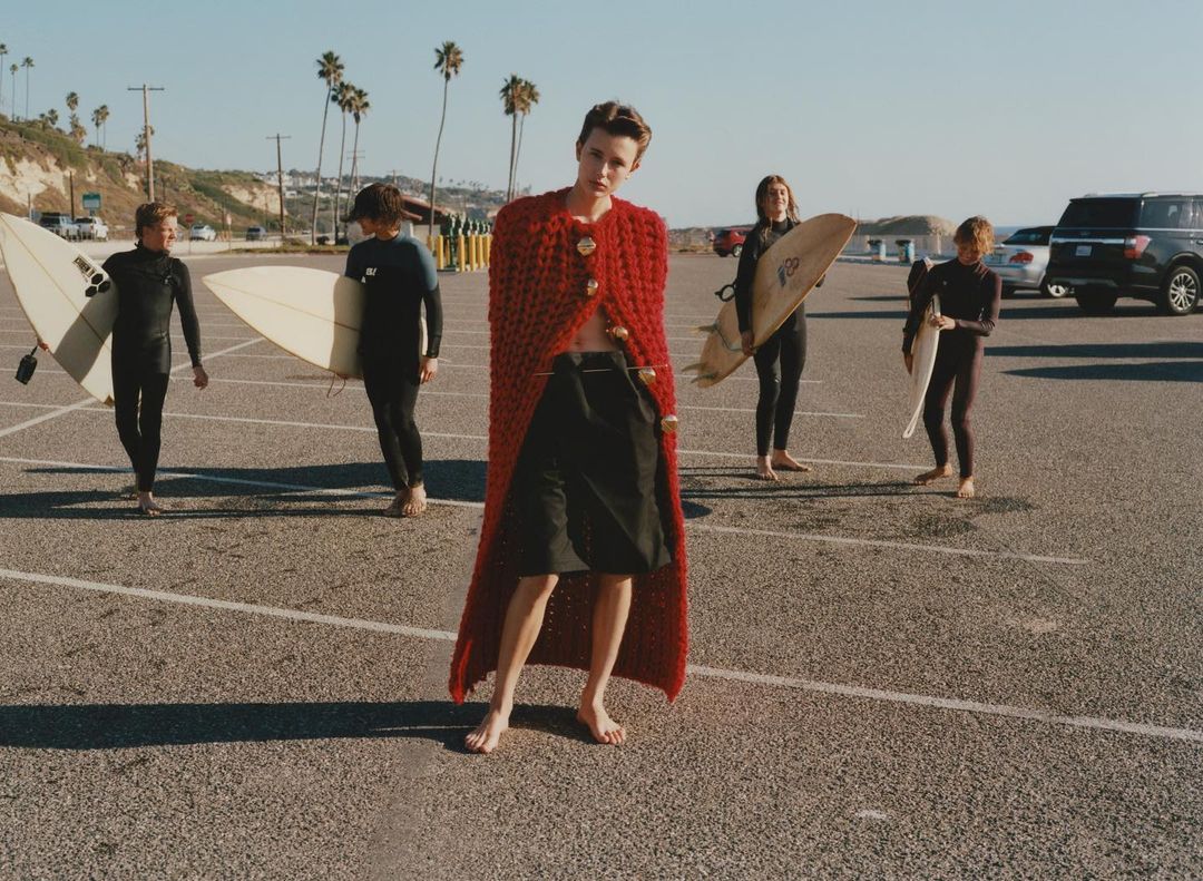 Fashionable person in red knit cape posing in a parking lot near the beach with a group of surfers holding surfboards.