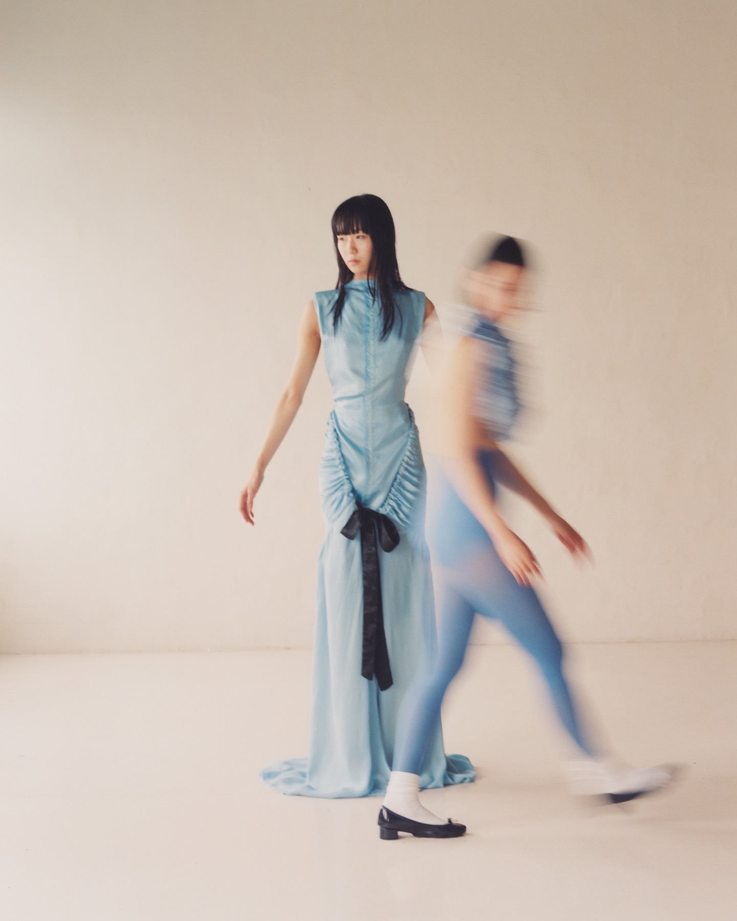 Two women pose in a studio setting, one in focus in a blue dress, the other blurred with motion, also in blue.
