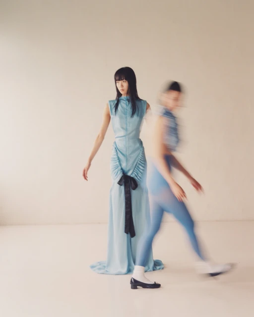 Two women pose in a studio setting, one in focus in a blue dress, the other blurred with motion, also in blue.