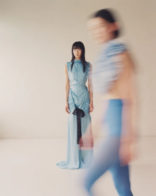 Two young Asian women pose in a light studio setting. One stands in focus wearing a blue dress with a black bow. Soft colors.
